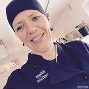 Köchin in professioneller Küche trägt schwarze Kochmütze und -jacke mit 'tophill kitchen' Logo, bereit für handgemachte kulinarische Kreationen, moderne Küchenausstattung im Hintergrund.