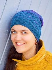 Bunte handgemachte Wollmütze mit Muster, getragen von einer lächelnden Frau, die an einer blauen Wand lehnt, ideal für Wintermode und handgefertigte Accessoires.