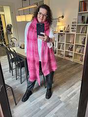 Frau mit Brille und pinkem Schal, die ein Selfie in einem Bücherregal macht, in einem gemütlichen Raum mit moderner Einrichtung.