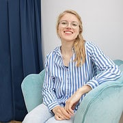 Junge Frau mit Brille und langem blondem Haar, lächelnd, in blauer gestreifter Bluse, sitzt entspannt auf einem Sessel in einem modernen Raum, passend für Handmade Kultur.