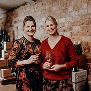 Zwei Frauen beim Weinverkosten, lachen und genießen in einer gemütlichen Atmosphäre mit Backsteinwand im Hintergrund.
