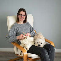 Händische Strickarbeiten, Frau beim Stricken mit Hund, gemütliche Handarbeit, kreative DIY-Projekte, Wohnzimmer-Atmosphäre, handmade Kultur, kreatives Hobby, kuschelige Wolle.