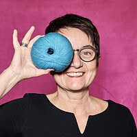 Buntes Porträt einer Frau mit Kurzhaarfrisur und Brille, die ein blaues Garnball hält, vor pinkem Hintergrund, für Handmade Kultur, kreative Handarbeit, Stricken, Häkeln, DIY, Handarbeiten, Nähen.