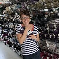 Bündel schwarzer Wolle in den Armen einer lachenden Frau, die in einem kreativen Handarbeitsgeschäft mit vielen Wollspulen im Hintergrund steht.