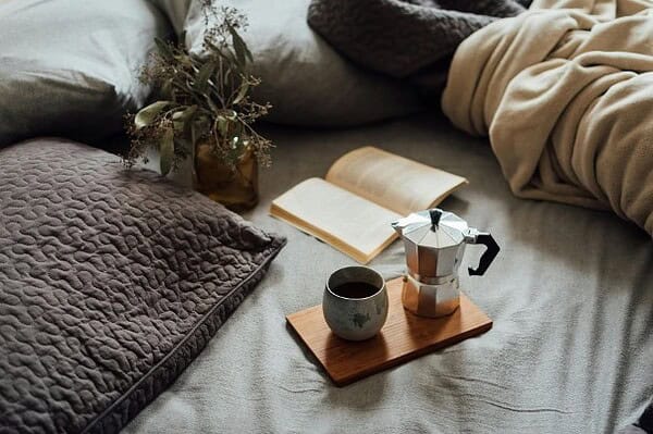 Håndgemachte Kultur – Gemütliches Bett mit Kaffeetasse, Mokka, Buch und Pflanzen, entspannte Atmosphäre, perfekte Kulisse für Handwerkskunst und kreative Momente.