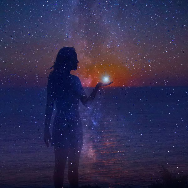 Frau hält leuchtenden Stern im Hand, umgeben von Sternen und Milchstraße, bei Sonnenuntergang am Meer, magische und ruhige Atmosphäre, ideal für Entspannung und Selbstfürsorge.